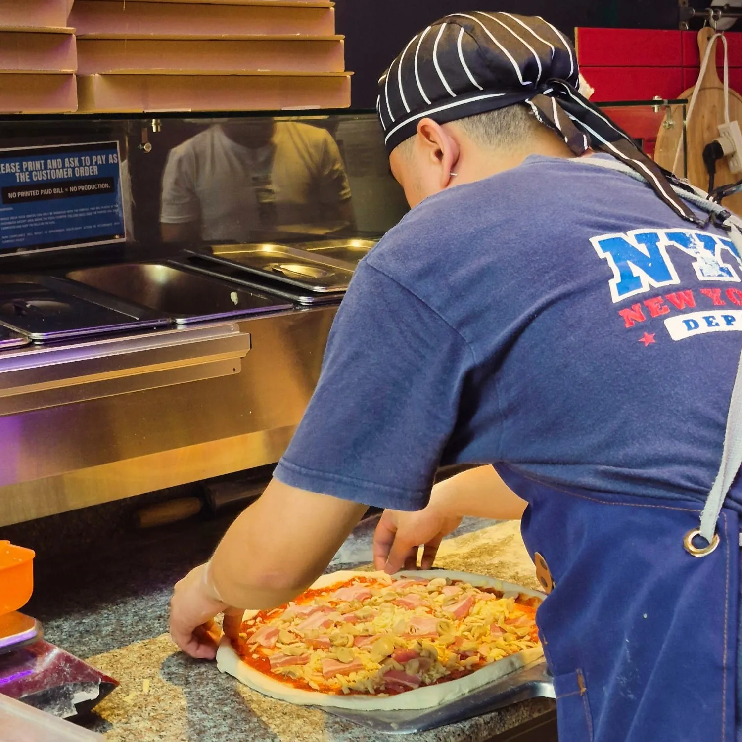 A NYPD Pizza 99 chef in a New York Department apron shaping a pizza behind the counter.
